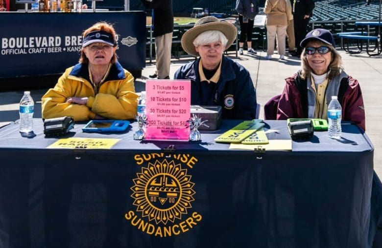 Three women sitting at a table with a sign.