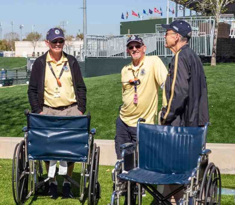 Three men standing next to two wheel chairs.